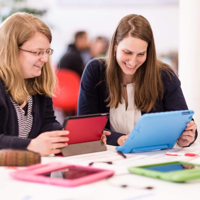 Zwei Frauen sitzen an einem Tisch mit Tablets und halten je eines in den Händen.