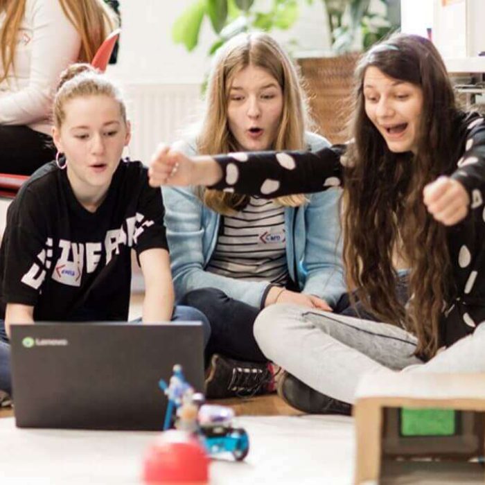 Drei Mädchen sitzen vor einem Computer und freuen sich, im Vordergrund ein Roboter.