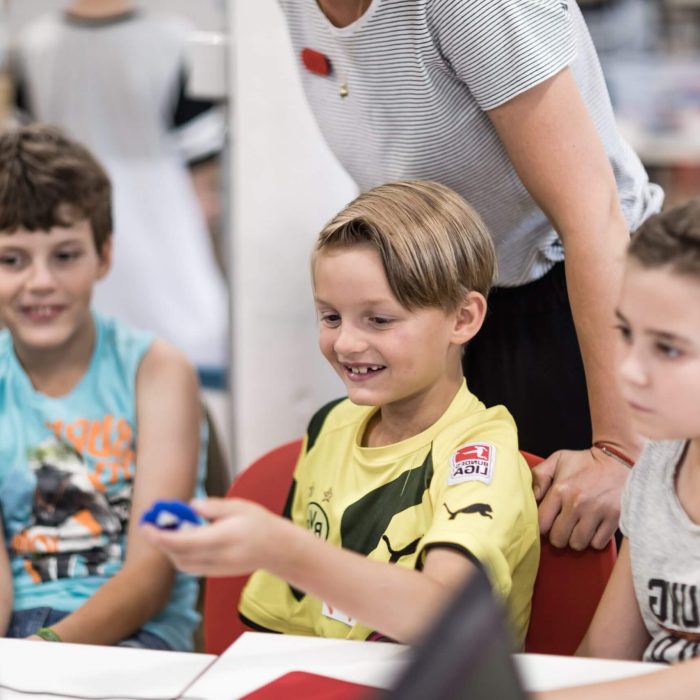 Drei lächelnde Kinder sitzen an einem Tisch, ein blonder Junge in der Mitte hält etwas blaues in der Hand. Im Hintergrund steht eine Frau, zu sehen ist nur der Oberkörper.