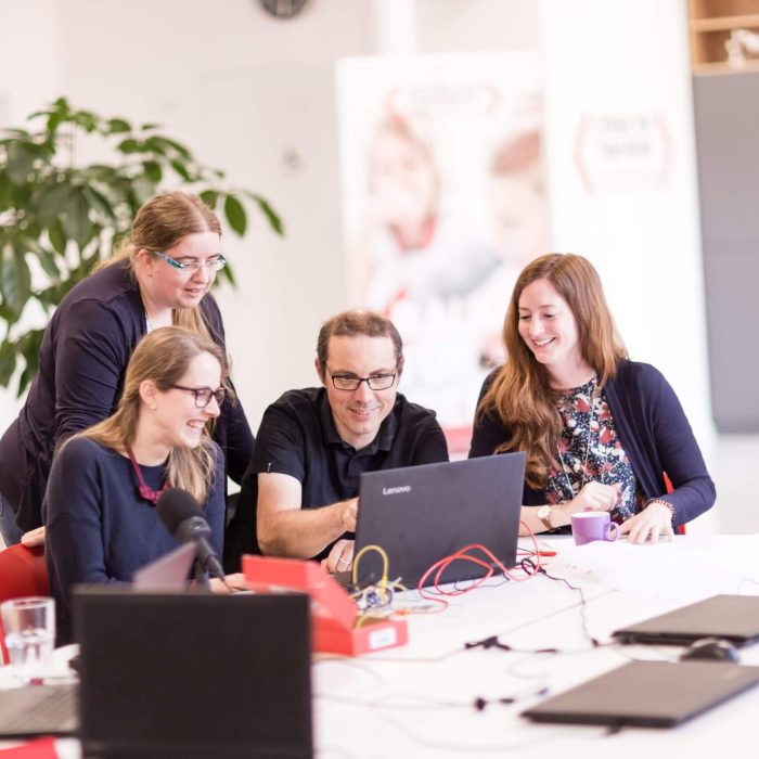 Drei sitzende Personen und eine stehende Frau schauen auf einen Laptop. Auf dem Tisch liegen mehrere Laptops und Kabel.