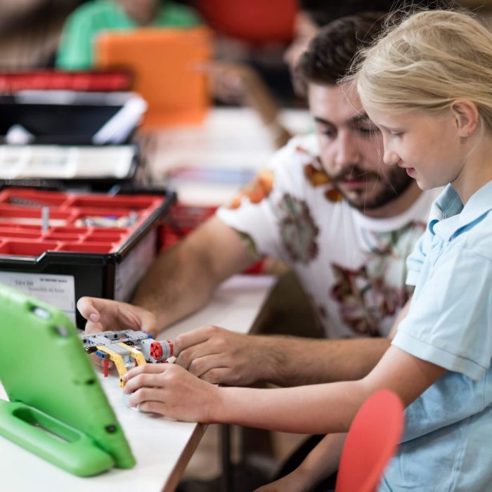 Ein blondes Mädchen und ein Mann sitzen an einem Schreibtisch und bauen einen Lego-Roboter zusammen. Auf dem Schreibtisch stehen Legokisten und IPads.