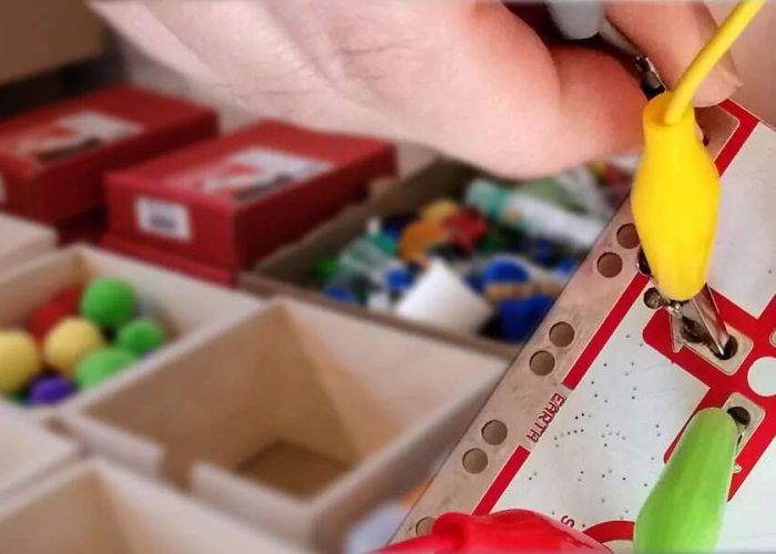 Hand hält eine Leiterplatine mit bunten Kabeln, im Hintergrund ist Bastelmaterial zu sehen.